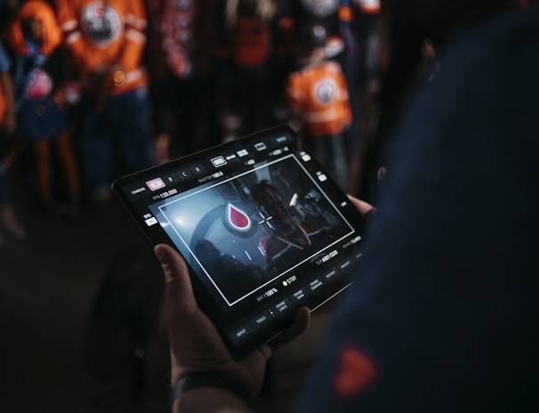 A person is holding a tablet that displays a video or image of a red and blue emblem on the screen. In the background, several people wearing dark and orange colored clothing, possibly sports jerseys, are partially visible. The scene is dimly lit, suggesting an indoor or evening setting.
