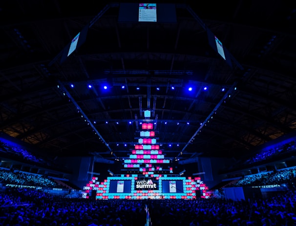 An expansive venue filled with a large audience, featuring a grand stage lit with vibrant blue and pink lights. The ceiling is decorated with a large display, and numerous screens showcase the event's branding. The atmosphere is dynamic and futuristic, reflecting a technologically advanced conference.